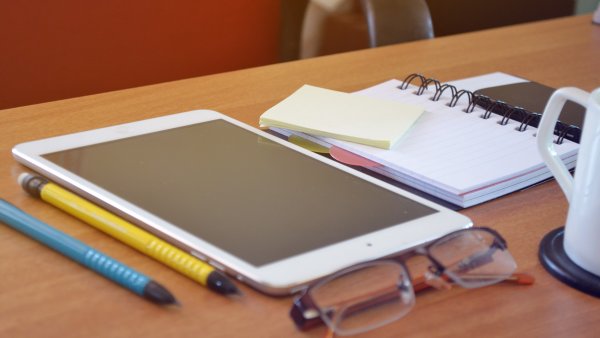 Auf einer Tischplatte liegen zwei Stifte, eine Brille, ein Tablet und ein Notizbuch