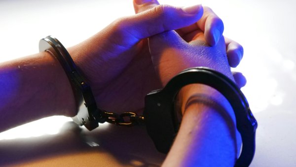 Hände sind in Handschellen und liegen auf einem weißen Tisch, blaues Licht beleuchtet die Szene
