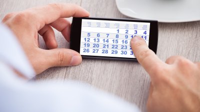 Eine Hand hält ein Smartphone mit einem Kalender auf dem Bildschirm, mit einem Zeigefinder auf dem Display 